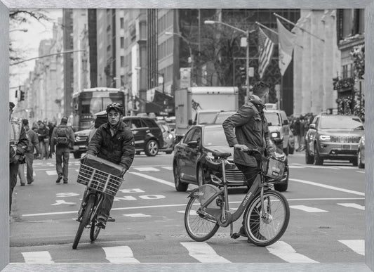 A black and white street photograph of a busy city intersection, framed in silver. In the foreground, two men are crossing with their bicycles amidst traffic, with one cyclist looking directly at the camera. Tall buildings and other pedestrians line the background, capturing a moment of everyday city life. Artwork