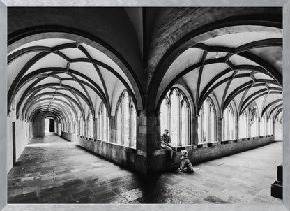 A black and white photograph of a person and their dog sitting in the corner of a Gothic cloister. The stone hallway features a dramatic vaulted ceiling and a long row of arched windows, creating a powerful sense of perspective and light. Decor