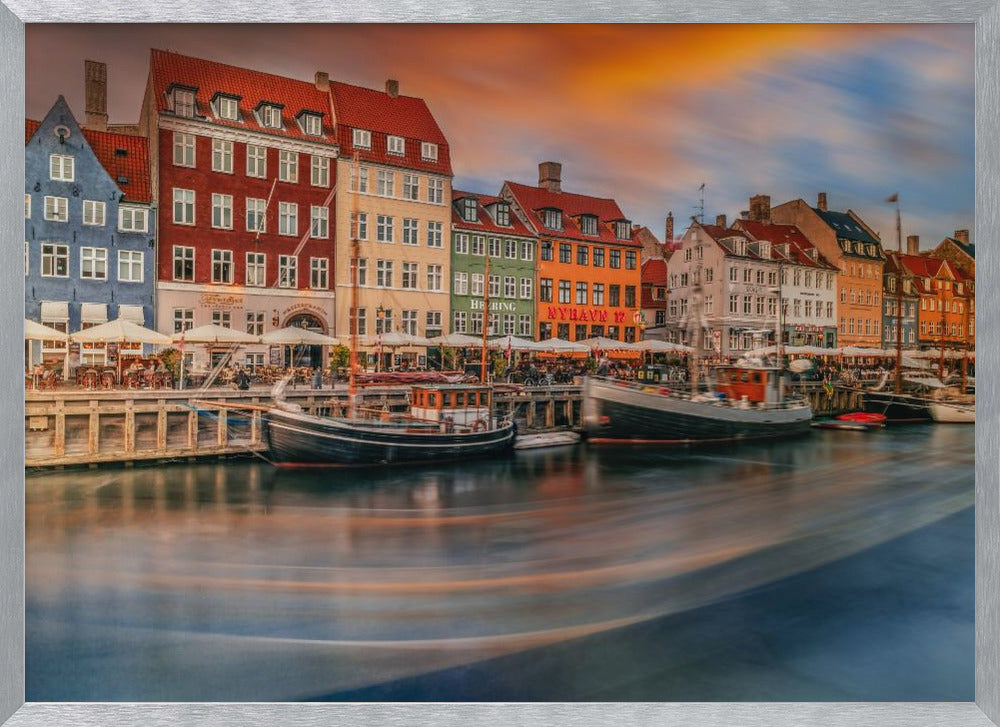 A long-exposure photograph of the colorful Nyhavn harbor in Copenhagen at sunset. Historic, brightly colored buildings line the canal, where two traditional wooden boats are moored. The sky is a dramatic blend of orange and blue, and the water is blurred, showing streaks of reflected light. Decor