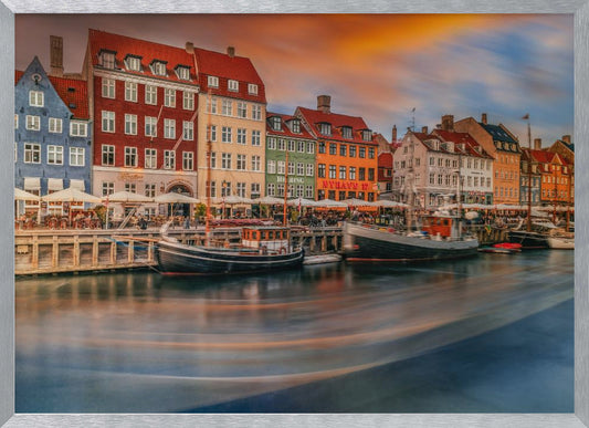 A long-exposure photograph of the colorful Nyhavn harbor in Copenhagen at sunset. Historic, brightly colored buildings line the canal, where two traditional wooden boats are moored. The sky is a dramatic blend of orange and blue, and the water is blurred, showing streaks of reflected light. Decor