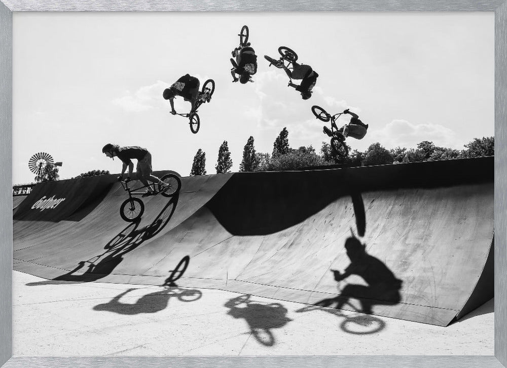 A dynamic black and white action shot showing the progression of a BMX rider doing a backflip off a large ramp. Multiple exposures capture the rider at different points in the air, creating an arc of motion against a bright sky. Strong shadows are cast on the ramp below. Print