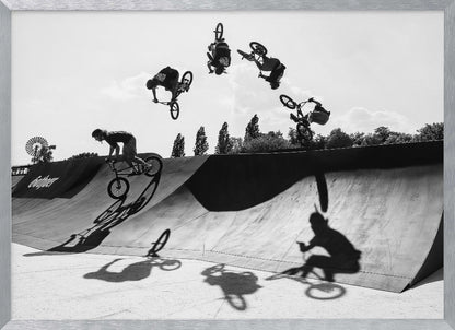 A dynamic black and white action shot showing the progression of a BMX rider doing a backflip off a large ramp. Multiple exposures capture the rider at different points in the air, creating an arc of motion against a bright sky. Strong shadows are cast on the ramp below. Print