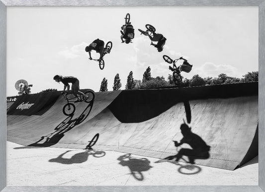 A dynamic black and white action shot showing the progression of a BMX rider doing a backflip off a large ramp. Multiple exposures capture the rider at different points in the air, creating an arc of motion against a bright sky. Strong shadows are cast on the ramp below. Print