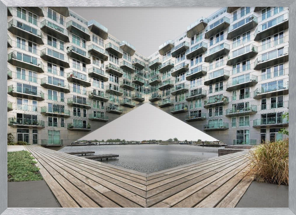 A symmetrical, mirrored photograph of a modern V-shaped apartment complex with numerous glass balconies, overlooking a wooden deck and a calm body of water under a grey sky. The image is presented within a silver picture frame. Artwork