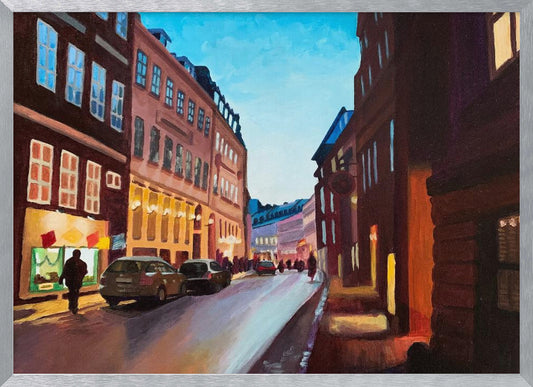 An impressionistic painting of a European city street at dusk, shown in a silver frame. The sky is a bright, clear blue, contrasting with the deep brown and maroon buildings. Warm yellow light glows from shop windows and street lamps, reflecting on the wet-looking street where cars are parked. Decor