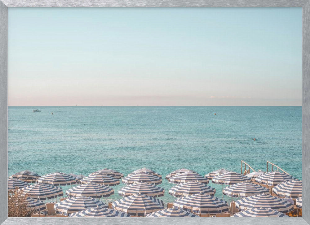 A high-angle photograph of a beach packed with blue and white striped umbrellas, with the turquoise ocean stretching out to a pale blue sky in the background. The entire image is enclosed in a silver frame. Poster