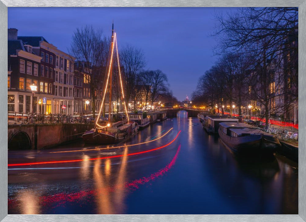 A long-exposure photograph of an Amsterdam canal at dusk. The scene is bathed in a deep blue light, with warm yellow and orange lights from street lamps and buildings reflecting on the water. A sailboat is decorated with glowing string lights, and red and white light trails from a passing boat create streaks across the canal. Houseboats are moored along the sides, and a bridge is visible in the distance. Poster