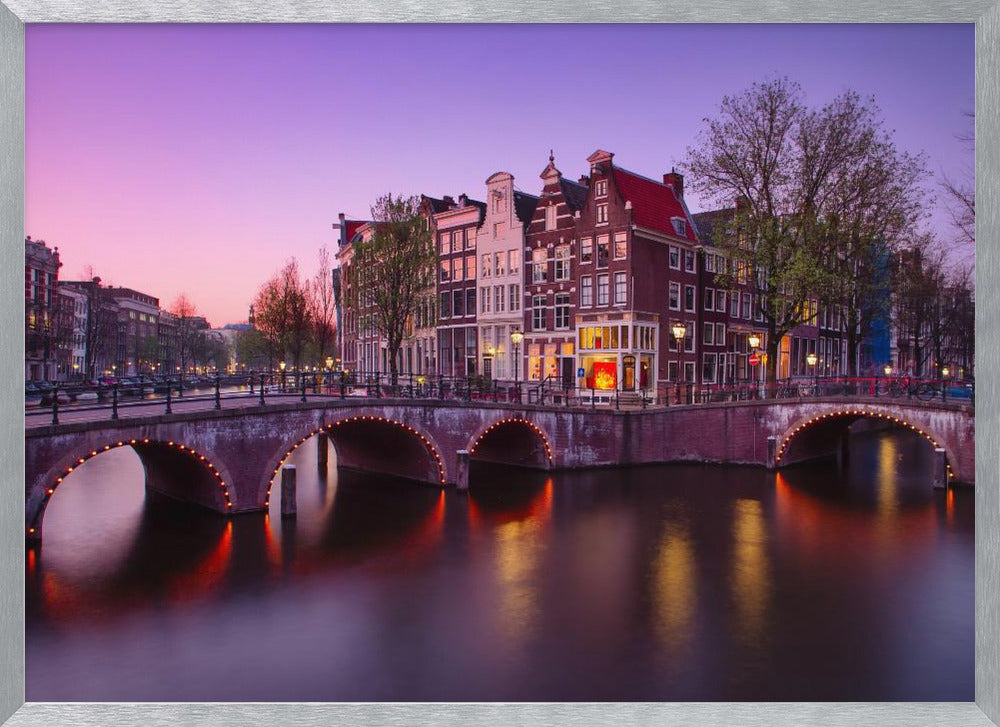 A scenic view of an Amsterdam canal at twilight, with historic brick buildings and illuminated arched bridges reflected in the calm water under a vibrant pink and purple sky. Poster