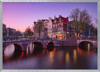 A scenic view of an Amsterdam canal at twilight, with historic brick buildings and illuminated arched bridges reflected in the calm water under a vibrant pink and purple sky. Poster