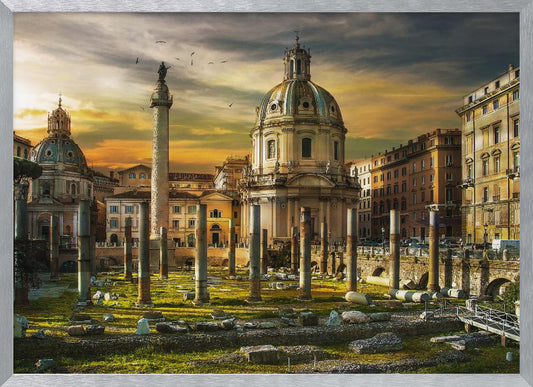 A framed photograph of Trajan's Forum in Rome, Italy, during a dramatic sunset. The foreground is filled with ancient stone columns and ruins on a grassy field, leading to the prominent Trajan's Column and the domed Church of the Most Holy Name of Mary against a golden and dark cloudy sky. Wall Art