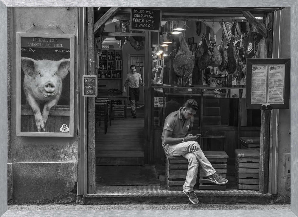 A framed, black and white street photograph of an Italian prosciutto shop. A young man sits on a wooden crate outside, looking at his phone, while cured hams hang inside the shop and a large, framed picture of a pig is on the exterior wall. Artwork