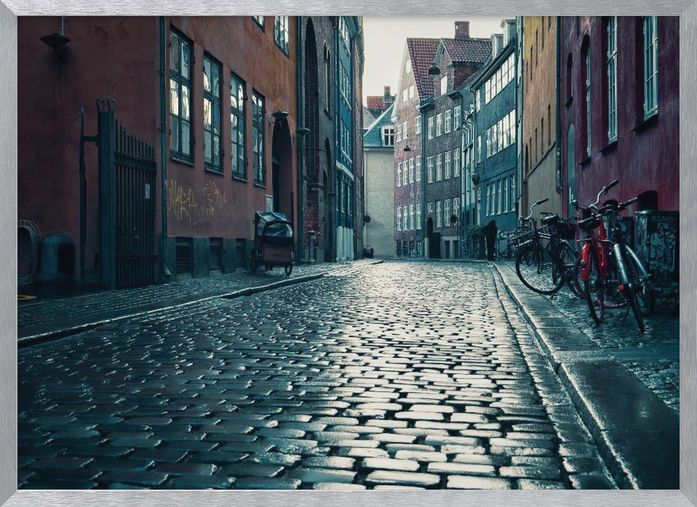 A low-angle shot of a wet, reflective cobblestone street in a narrow European alley. The street is lined with historic, colorful buildings in shades of red, orange, and blue under an overcast sky. Bicycles are parked along the sidewalk on the right, adding to the quiet, moody city scene. Poster