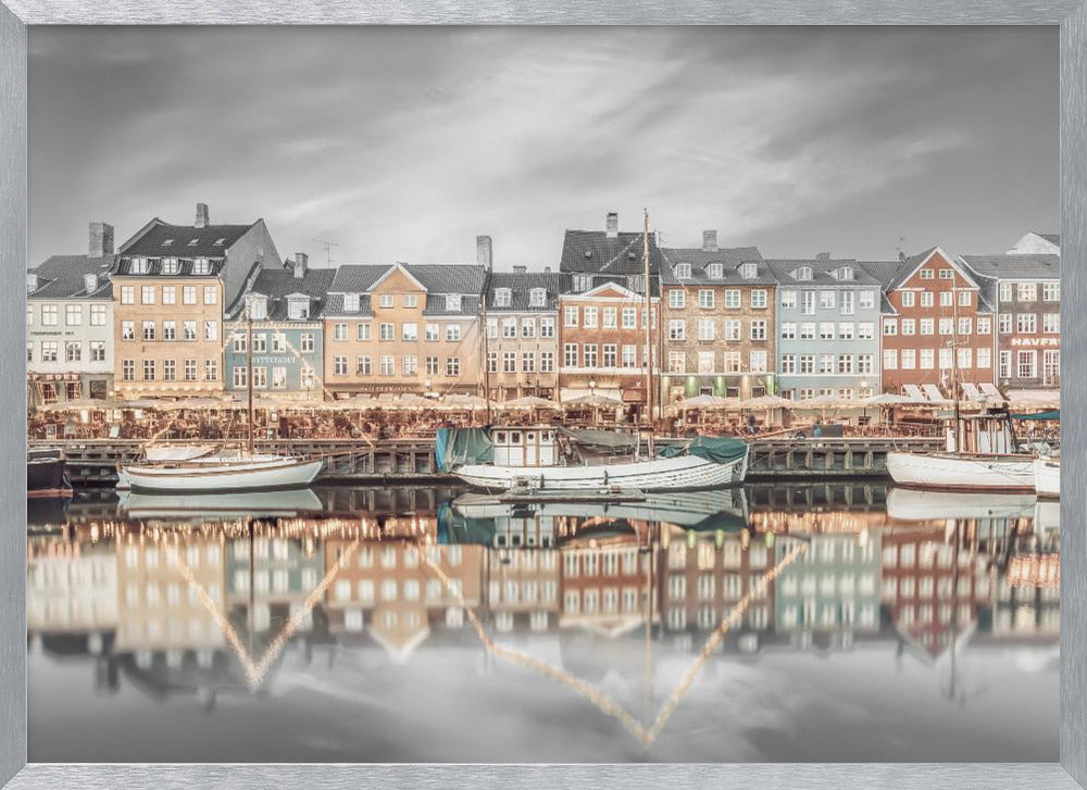 A framed, color-selective photograph of the Nyhavn harbor in Copenhagen. A row of colorful 17th and 18th-century townhouses lines the waterfront, with their warm lights and facades reflecting in the calm canal. The sky and parts of the water are in black and white, making the colored buildings stand out. Several boats are moored along the quay. Decor