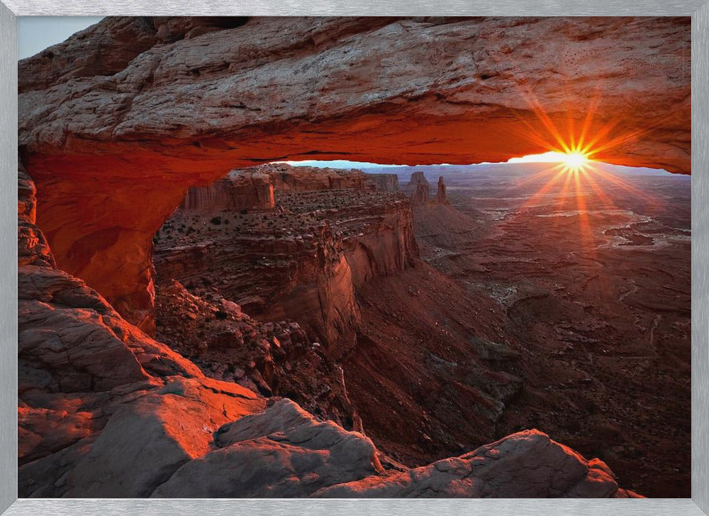 A spectacular sunrise seen through the natural frame of Mesa Arch in Canyonlands National Park, Utah. The sun bursts over the horizon, casting a dramatic orange and red glow on the underside of the rock arch and the vast canyon landscape below. Poster