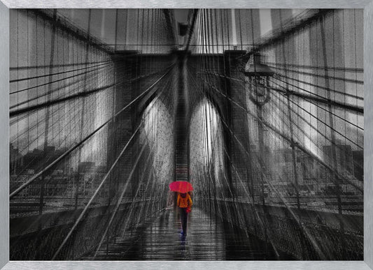 A motion-blurred, black and white photo of the Brooklyn Bridge with a person holding a bright red umbrella walking down the center. The selective color makes the red umbrella the focal point against the dramatic, almost abstract lines of the bridge's cables and arches on a wet, reflective walkway. The image is presented in a silver frame. Wall Art