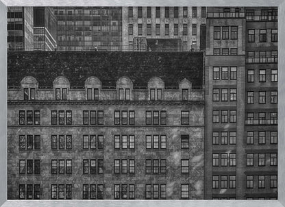 A dense, black and white photograph of various city buildings packed closely together. The image highlights the contrasting architectural styles, from an older stone building with a dark roof in the foreground to taller brick and modern glass buildings in the background, creating a geometric pattern of windows and textures. Decor