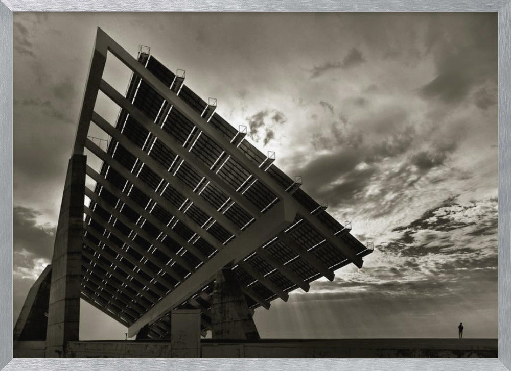 A framed, dramatic black and white photograph showing a massive, tilted solar panel structure from a low angle. The sky is filled with dark, moody clouds with some sun rays breaking through. A tiny silhouette of a person in the lower right corner emphasizes the grand scale of the architecture. Wall Art