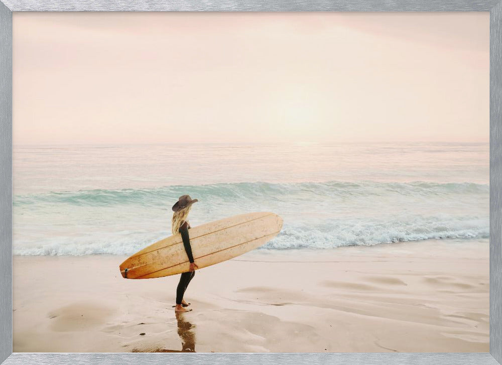 A female surfer with long blonde hair, wearing a black wetsuit and a hat, holds a surfboard while standing on a wet sandy beach looking out at the ocean during a hazy pink sunset. Decor