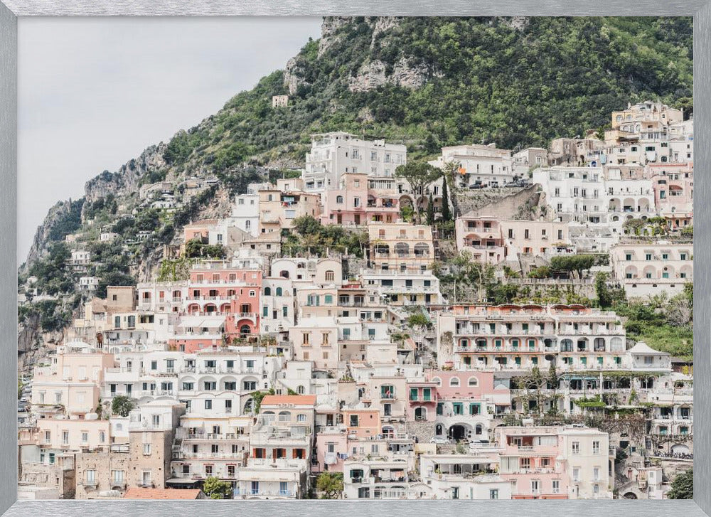 Photograph of the iconic cliffside village of Positano on the Amalfi Coast, showcasing a dense cascade of white, pink, and peach buildings down a lush green mountain under an overcast sky. The image is presented within a silver picture frame. Artwork