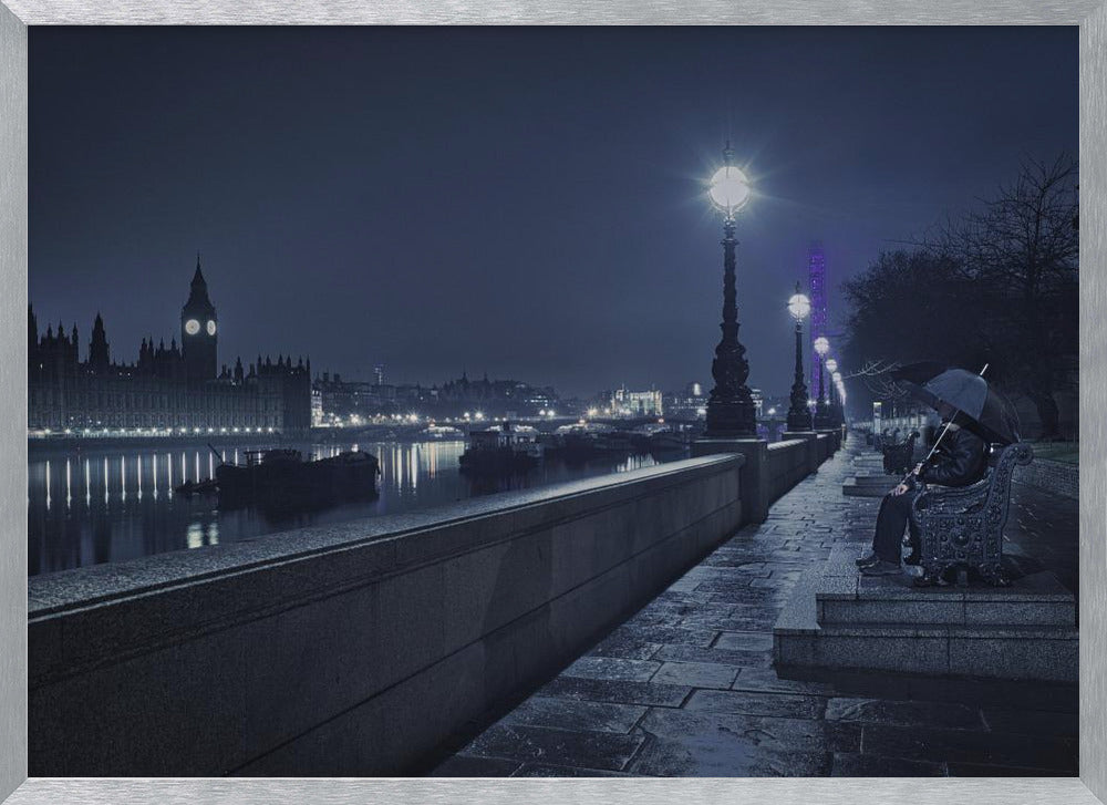 A moody, blue-toned night photograph of London. A solitary figure sits on a bench under an umbrella on a wet stone walkway along the River Thames, illuminated by vintage streetlights. Across the river, the iconic Houses of Parliament and Big Ben are lit up against the dark sky, their lights reflecting in the water. Poster