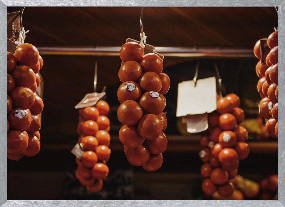A close-up, warm-toned photograph of several bunches of ripe red tomatoes hanging from hooks in a row, likely at a market or in a pantry. The background is dark and out of focus, highlighting the vibrant produce. Poster