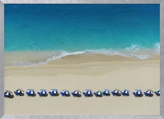 An aerial view of a pristine sandy beach with vibrant turquoise ocean waves gently lapping the shore. A long, neat row of blue and white striped beach umbrellas stands along the sand, creating a serene and orderly coastal scene, all enclosed within a silver frame. Artwork