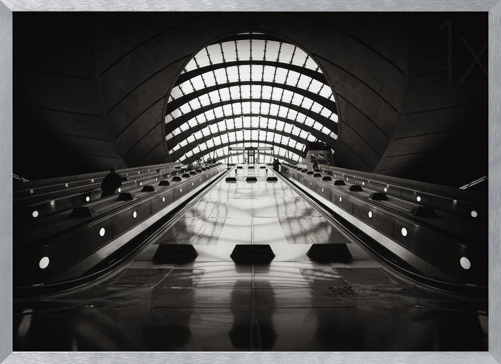 A dramatic low-angle, black and white photograph of the escalators at Canary Wharf station. The perspective looks up the sleek, reflective escalators towards a massive, arched, grid-patterned window at the far end, creating strong leading lines and a sense of scale within the modern, curved architecture. Decor