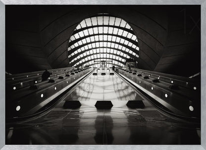 A dramatic low-angle, black and white photograph of the escalators at Canary Wharf station. The perspective looks up the sleek, reflective escalators towards a massive, arched, grid-patterned window at the far end, creating strong leading lines and a sense of scale within the modern, curved architecture. Decor