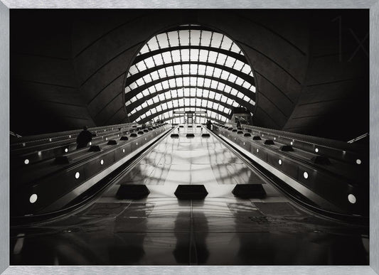 A dramatic low-angle, black and white photograph of the escalators at Canary Wharf station. The perspective looks up the sleek, reflective escalators towards a massive, arched, grid-patterned window at the far end, creating strong leading lines and a sense of scale within the modern, curved architecture. Decor