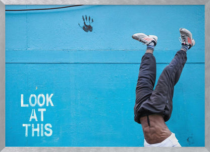 A person's lower body doing a handstand against a bright blue wall with the stenciled words 'LOOK AT THIS' in white. The person wears dark pants and sneakers, and their muscular abdomen is visible. A black handprint is on the wall above the text. The artwork is presented in a silver frame. Print
