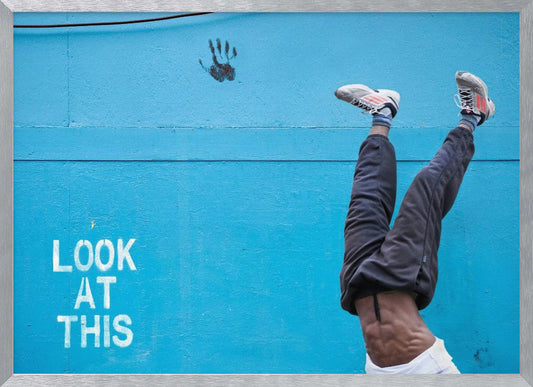 A person's lower body doing a handstand against a bright blue wall with the stenciled words 'LOOK AT THIS' in white. The person wears dark pants and sneakers, and their muscular abdomen is visible. A black handprint is on the wall above the text. The artwork is presented in a silver frame. Print