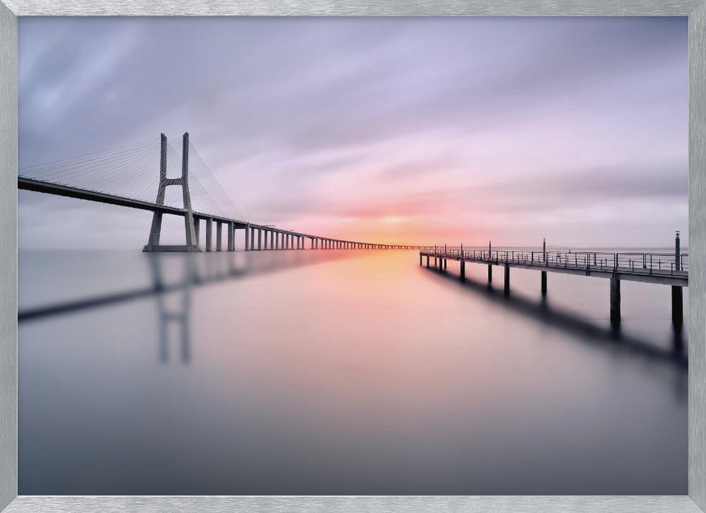 A serene long-exposure photograph of a modern cable-stayed bridge and a pier stretching over mirror-like water at sunset. The sky is filled with soft, streaky clouds in hues of pink and purple, reflected on the calm water's surface, all enclosed in a silver frame. Decor