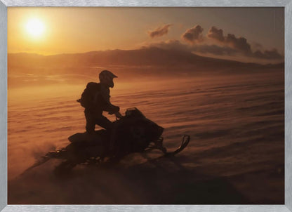 A silhouette of a person wearing a helmet and backpack stands on a snowmobile in a vast, snowy landscape during a golden sunset. The wind blows snow across the ground, creating a hazy, atmospheric effect, with mountains visible in the distance under a warm sky. Wall Art