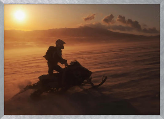 A silhouette of a person wearing a helmet and backpack stands on a snowmobile in a vast, snowy landscape during a golden sunset. The wind blows snow across the ground, creating a hazy, atmospheric effect, with mountains visible in the distance under a warm sky. Wall Art