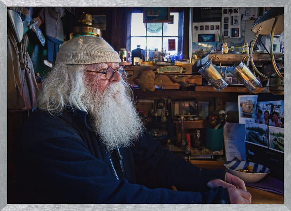 A profile portrait of an elderly man with a long, full white beard and hair, wearing glasses and a cream-colored knit beanie. He sits indoors in a warmly lit, cluttered room that looks like a cabin or a pub, with his hands clasped in front of him, gazing thoughtfully to the side. Poster