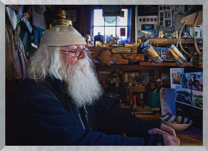 A profile portrait of an elderly man with a long, full white beard and hair, wearing glasses and a cream-colored knit beanie. He sits indoors in a warmly lit, cluttered room that looks like a cabin or a pub, with his hands clasped in front of him, gazing thoughtfully to the side. Poster