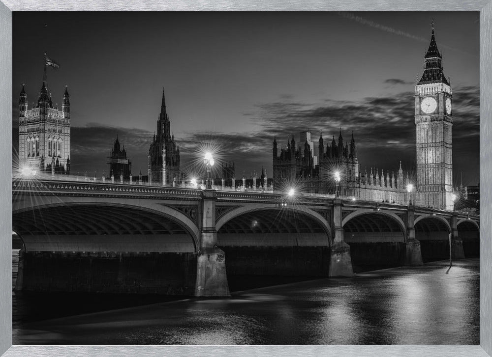A framed, black and white nighttime photograph of Westminster Bridge in London. The iconic Houses of Parliament and the glowing Big Ben clock tower stand prominently in the background against a dark, cloudy sky. The streetlights on the bridge create starburst effects, and their reflections shimmer on the surface of the River Thames in the foreground. Poster