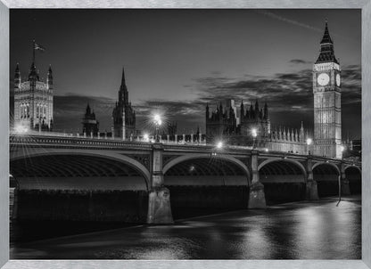 A framed, black and white nighttime photograph of Westminster Bridge in London. The iconic Houses of Parliament and the glowing Big Ben clock tower stand prominently in the background against a dark, cloudy sky. The streetlights on the bridge create starburst effects, and their reflections shimmer on the surface of the River Thames in the foreground. Poster