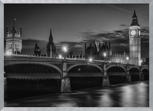 A framed, black and white nighttime photograph of Westminster Bridge in London. The iconic Houses of Parliament and the glowing Big Ben clock tower stand prominently in the background against a dark, cloudy sky. The streetlights on the bridge create starburst effects, and their reflections shimmer on the surface of the River Thames in the foreground. Poster
