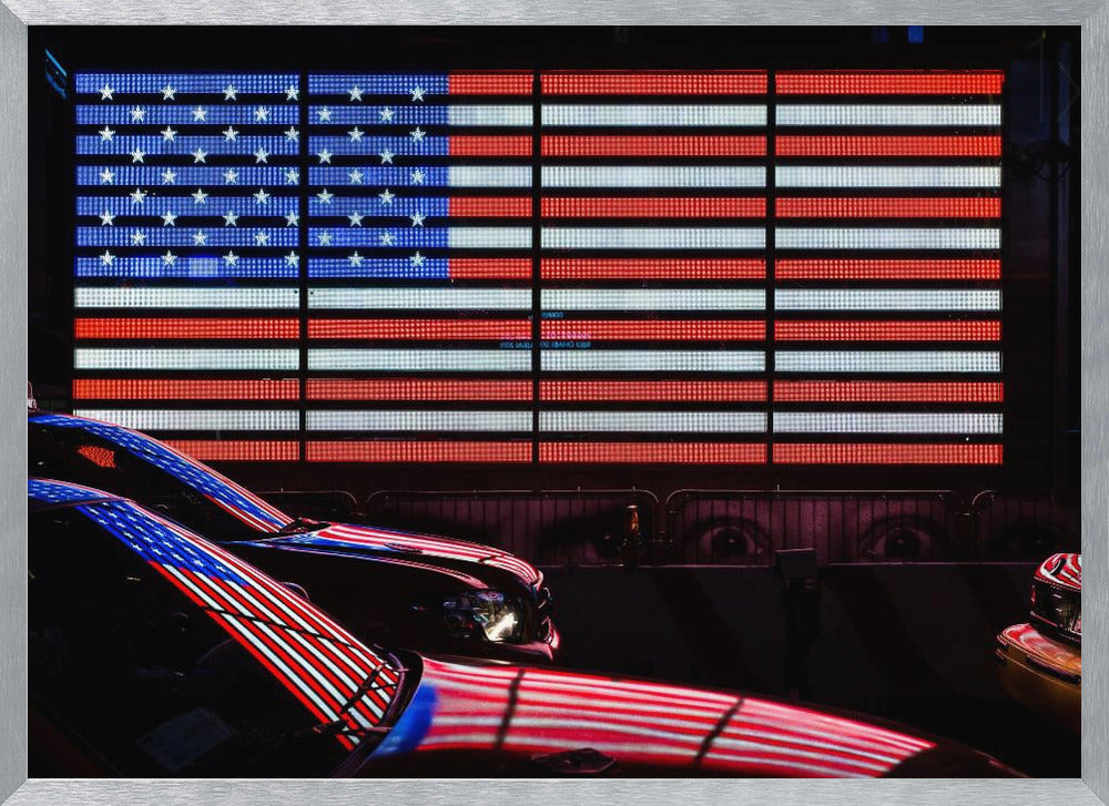 A large illuminated American flag on a billboard at night, with its red, white, and blue lights reflecting on the glossy hoods of two cars in the foreground. Wall Art