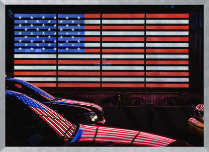 A large illuminated American flag on a billboard at night, with its red, white, and blue lights reflecting on the glossy hoods of two cars in the foreground. Wall Art