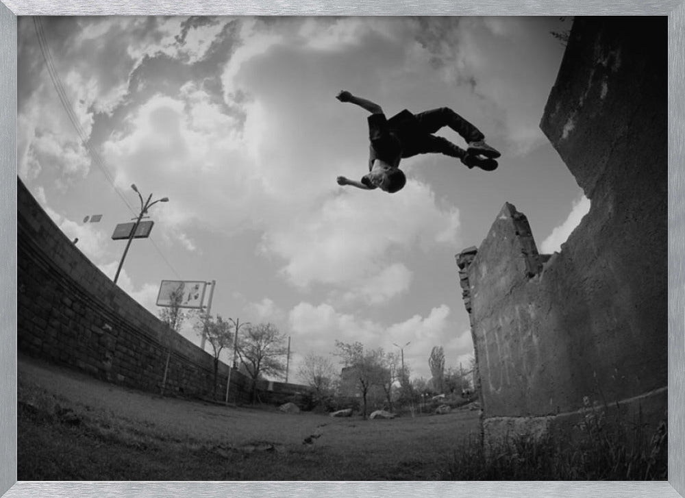A dynamic black and white fisheye photograph of a person mid-flip, jumping over a crumbling wall in an urban setting under a cloudy sky. The image has a silver-colored frame. Print