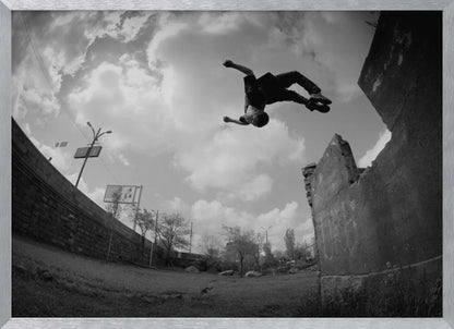 A dynamic black and white fisheye photograph of a person mid-flip, jumping over a crumbling wall in an urban setting under a cloudy sky. The image has a silver-colored frame. Print