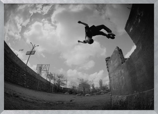 A dynamic black and white fisheye photograph of a person mid-flip, jumping over a crumbling wall in an urban setting under a cloudy sky. The image has a silver-colored frame. Print