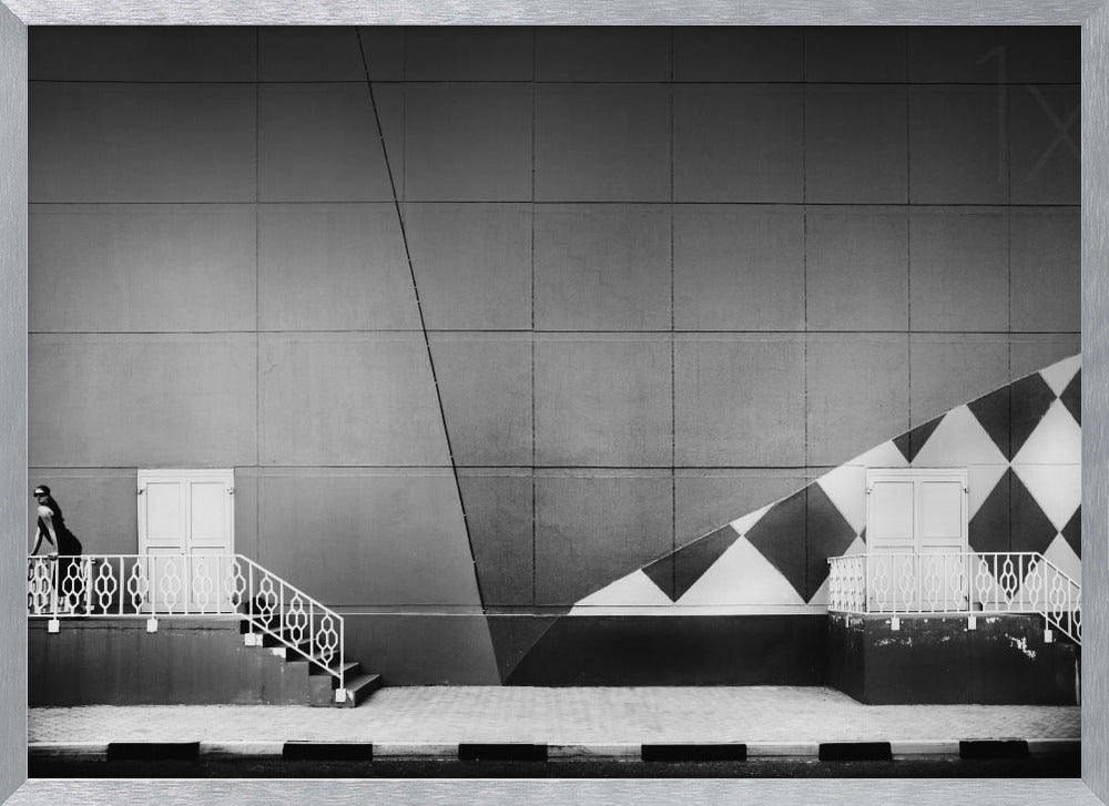 A framed black and white photograph of a modern urban building. A woman in a dark outfit stands on the left by a white ornate railing and door. The large gray wall behind her is bisected by strong diagonal lines and features a bold black and white diamond pattern on the right, where another door and railing are visible. Decor