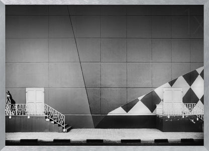 A framed black and white photograph of a modern urban building. A woman in a dark outfit stands on the left by a white ornate railing and door. The large gray wall behind her is bisected by strong diagonal lines and features a bold black and white diamond pattern on the right, where another door and railing are visible. Decor