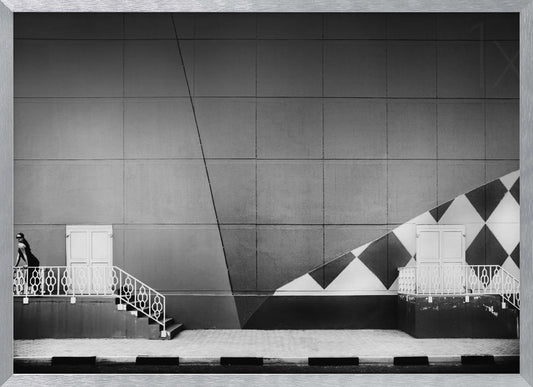 A framed black and white photograph of a modern urban building. A woman in a dark outfit stands on the left by a white ornate railing and door. The large gray wall behind her is bisected by strong diagonal lines and features a bold black and white diamond pattern on the right, where another door and railing are visible. Decor