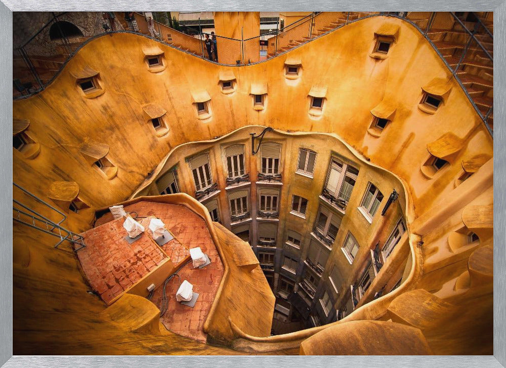 A high-angle photograph looking down into the surreal, undulating inner courtyard of an architectural masterpiece. The warm, golden-ochre walls curve organically, dotted with small, hooded windows, encircling the deep central lightwell that reveals floors of apartments below. Wall Art