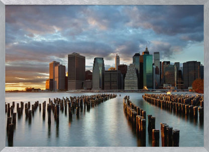 A framed photograph of the Lower Manhattan skyline at sunset, viewed from across the water. Rows of old wooden pier pilings lead the eye towards the city's skyscrapers, which are illuminated by the warm light of the setting sun under a dramatic, cloudy sky. Decor