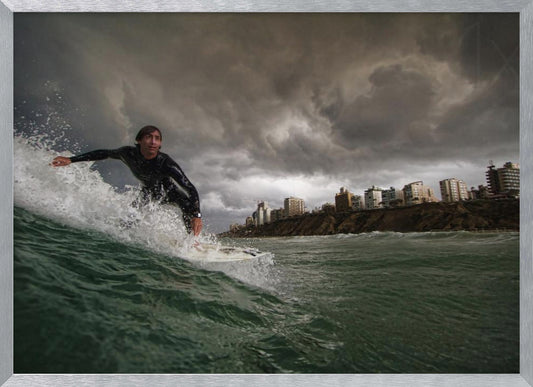 A close-up, low-angle shot of a male surfer in a black wetsuit riding a dark green ocean wave. In the background, a city skyline sits atop a cliff under a dramatic, dark, and stormy sky. The image is presented within a silver picture frame. Print